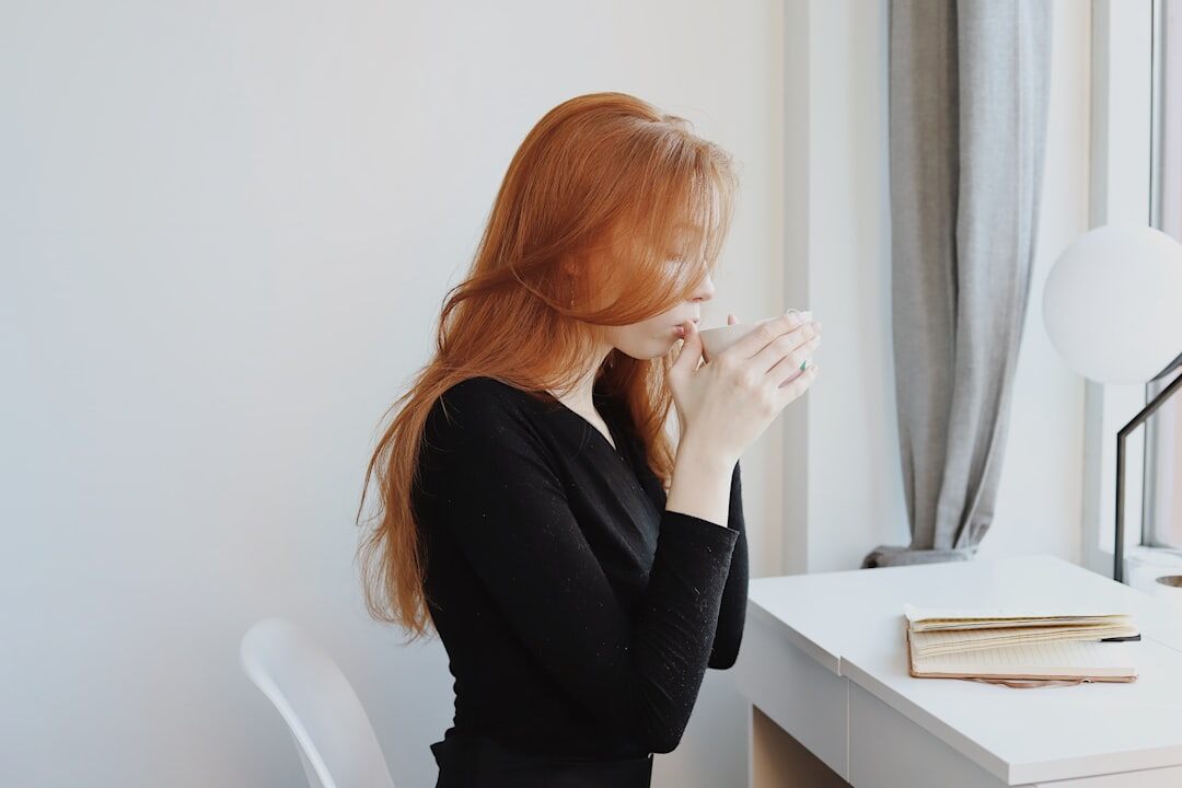 Woman writing thoughtfully in a journal at a wooden desk near a window